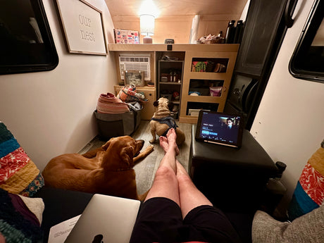 Cozy RV rental interior. Person's legs up, laptop & tablet (Three Pines playing), two dogs relaxing. "Our nest" sign above.