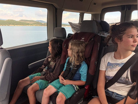 Three kids in an RV rental looking out at a large lake and distant hills on a scenic family road trip.