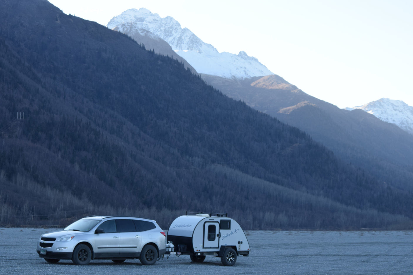 Silver SUV towing a Bushwhacker teardrop RV rental, parked among forested and snowy mountains.