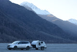 Silver SUV towing a Bushwhacker teardrop RV rental, parked among forested and snowy mountains.