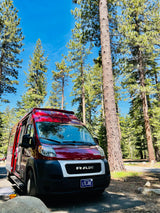 Red Ram ProMaster RV rental van parked in a sunny pine forest. The side door is open, inviting a closer look at the camping setup.