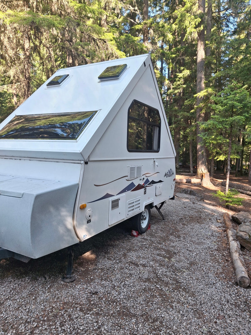  surrounded by tall trees.|White A-frame RV rental parked on a gravel campsite amidst tall forest trees.|White Chalet pop-up camper — photo 2