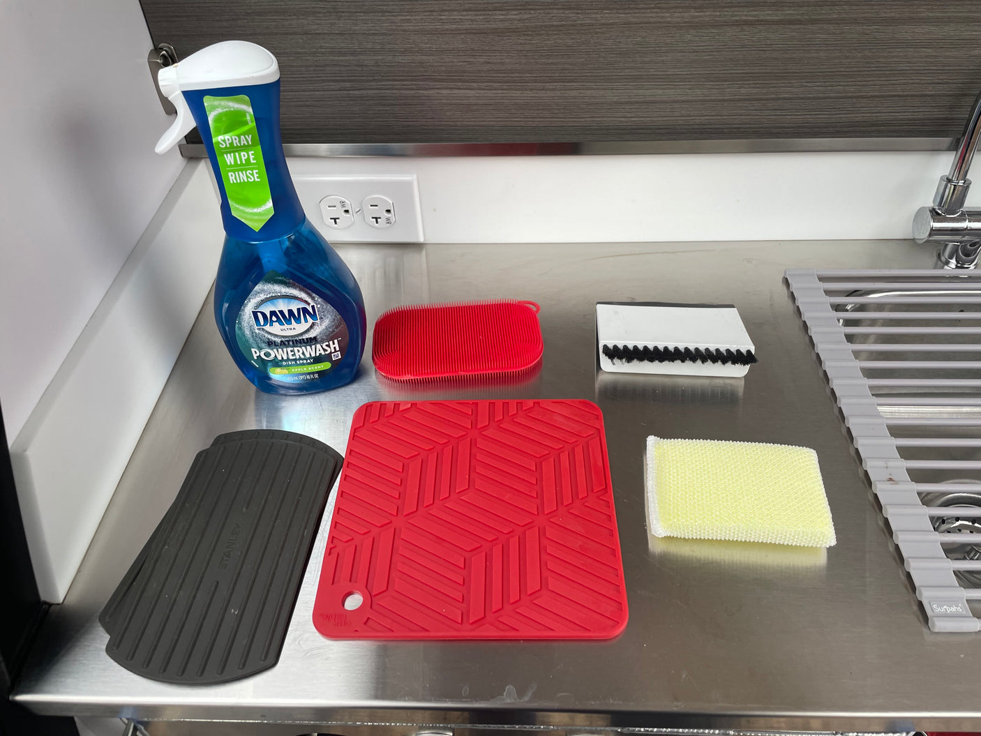 RV rental kitchen setup: Dawn Powerwash, sponges, brushes, silicone hot pads, and a roll-up drying rack on stainless steel.