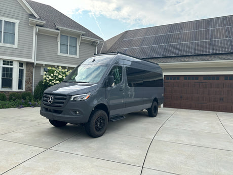 Modern grey Mercedes-Benz Sprinter van, ready for rv rental, parked on a concrete driveway in front of a house with solar panels.