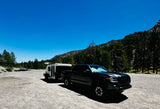 Black pickup truck towing a white teardrop RV rental on a gravel shoulder, sunny mountain road, blue sky & trees.
