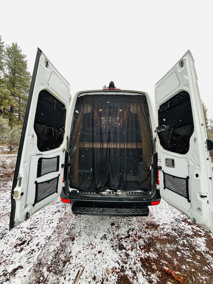 This RV rental van, with its rear doors open and a black mesh screen inside, is parked in a snowy forest setting.