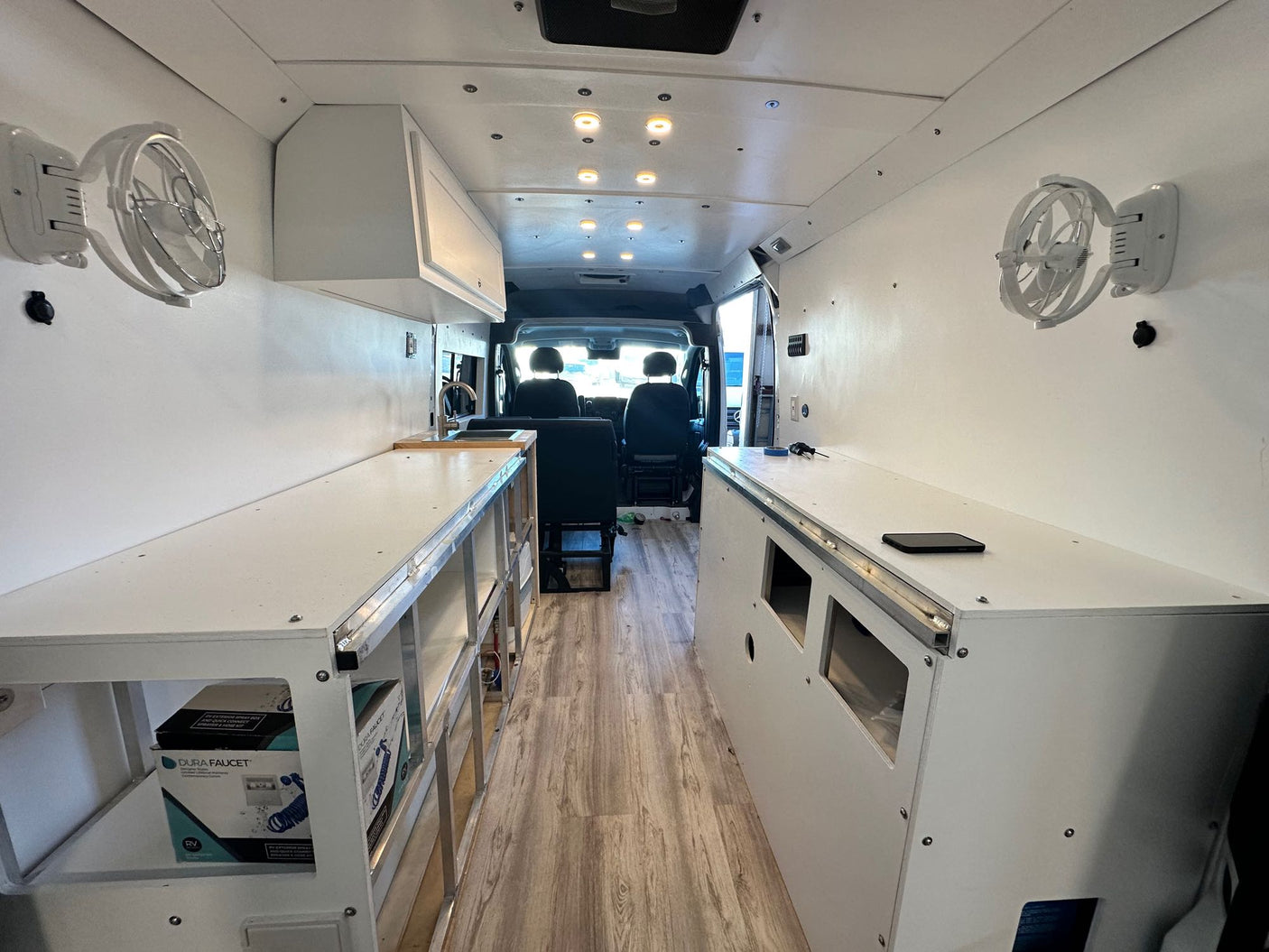 Bright RV rental interior: custom white van conversion with overhead lights, wall fans, clean counters, & wood-look floor.