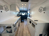 Bright RV rental interior: custom white van conversion with overhead lights, wall fans, clean counters, & wood-look floor.