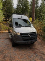 Silver Mercedes Sprinter RV rental parked on a gravel path with pine needles, surrounded by tall evergreen trees.