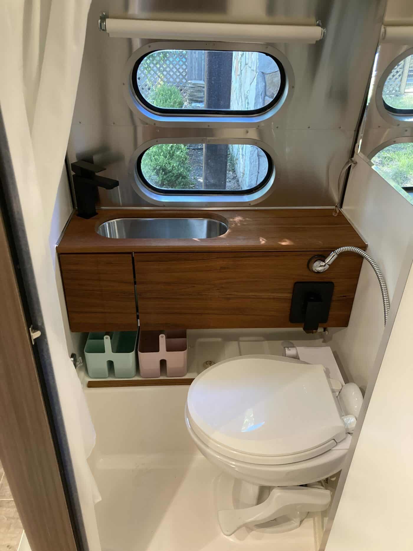 Inside a modern rv rental bathroom: wood vanity, sink, black faucet, toilet, handheld shower, and oval windows.