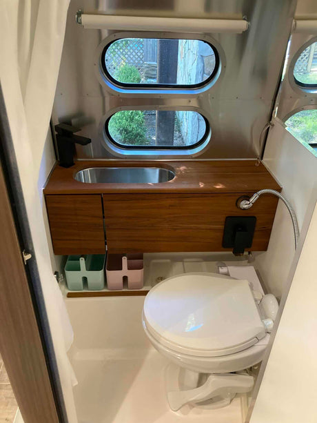 Inside a modern rv rental bathroom: wood vanity, sink, black faucet, toilet, handheld shower, and oval windows.