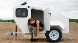 A woman sits in the open door of a white teardrop camper, tying shoes. Experience an RV rental!