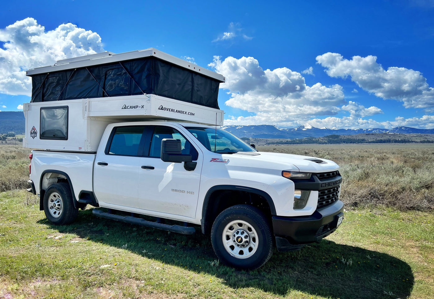 White Chevy truck with pop-top RV rental camper, parked in a grassy field with mountains under a blue sky. — photo 3