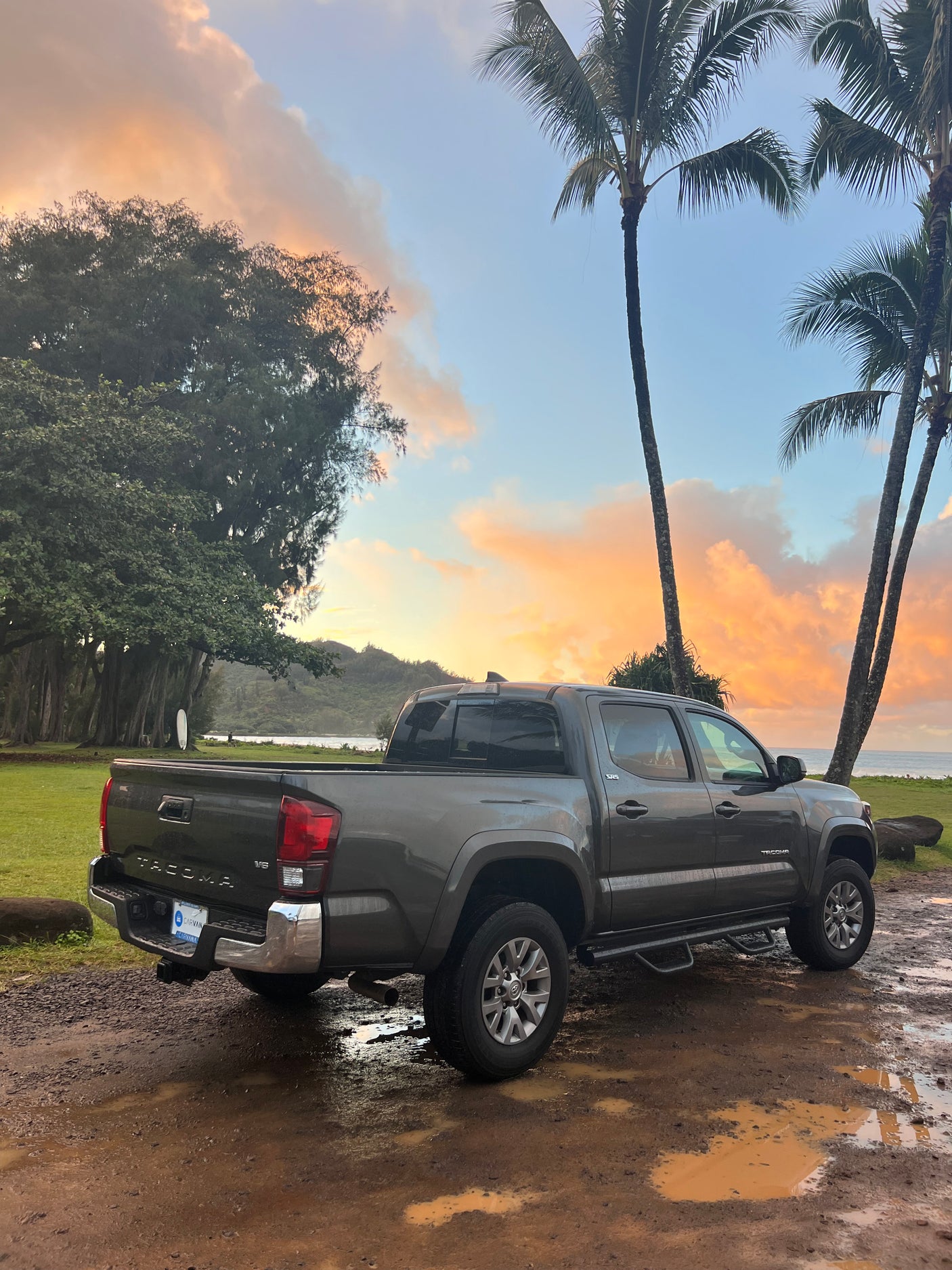 Gray Toyota Tacoma pickup on muddy path, beach sunset backdrop with palms. Great for adventure travel, like an RV rental.
