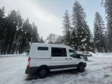 A white camper van (RV rental) is parked in a snowy lot, surrounded by tall pines, with Half Dome visible in the misty background.