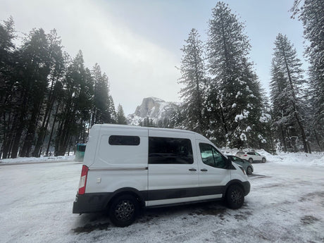 A white camper van (RV rental) is parked in a snowy lot, surrounded by tall pines, with Half Dome visible in the misty background.