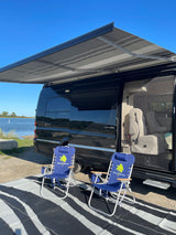 Black Airstream Interstate rv rental with awning. Two Tommy Bahama chairs & mat by a lake. Relaxing outdoor setup.