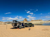 Explore! An off-road RV rental (green Jeep & overland trailer) set up on a sandy beach by a lake with desert cliffs. Ideal for adventure.