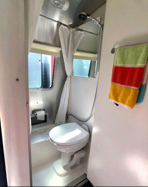 Inside an RV rental: a bright bathroom with toilet, shower, window, curtain, and striped towels hanging.