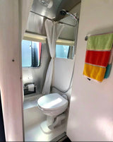 Inside an RV rental: a bright bathroom with toilet, shower, window, curtain, and striped towels hanging.