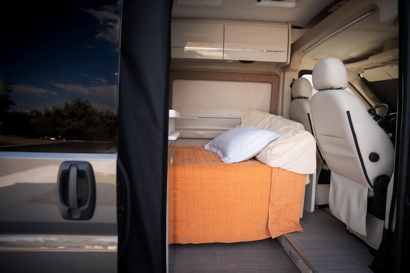 View inside a modern RV rental: a cozy bed with orange blanket, overhead storage, and white driver/passenger seats. — photo 19