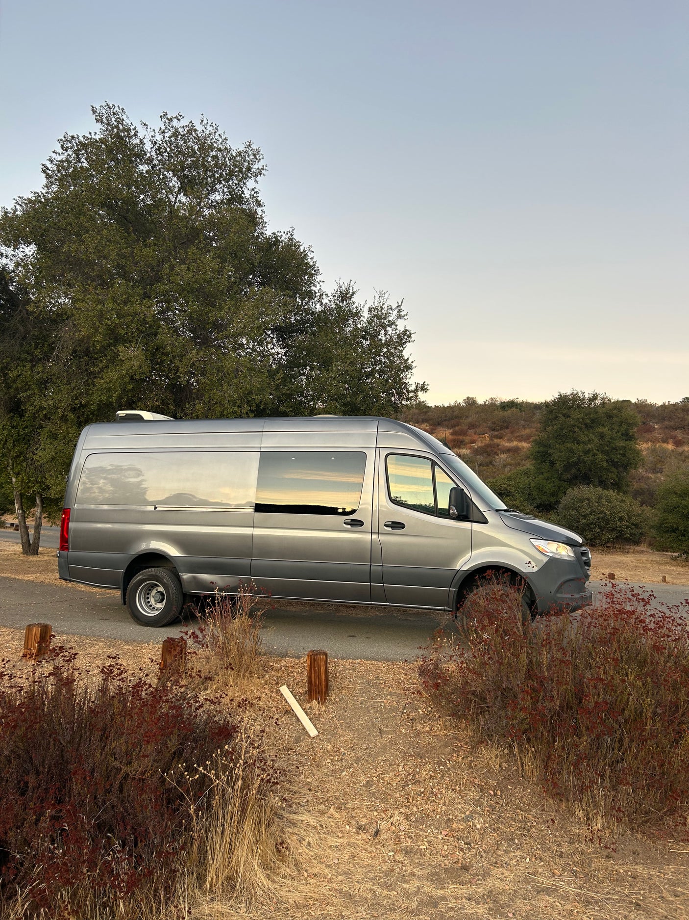 A gray Sprinter RV rental, parked on a paved road. Its windows reflect the golden hour light in a serene natural setting.