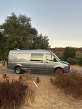 A gray Sprinter RV rental, parked on a paved road. Its windows reflect the golden hour light in a serene natural setting.