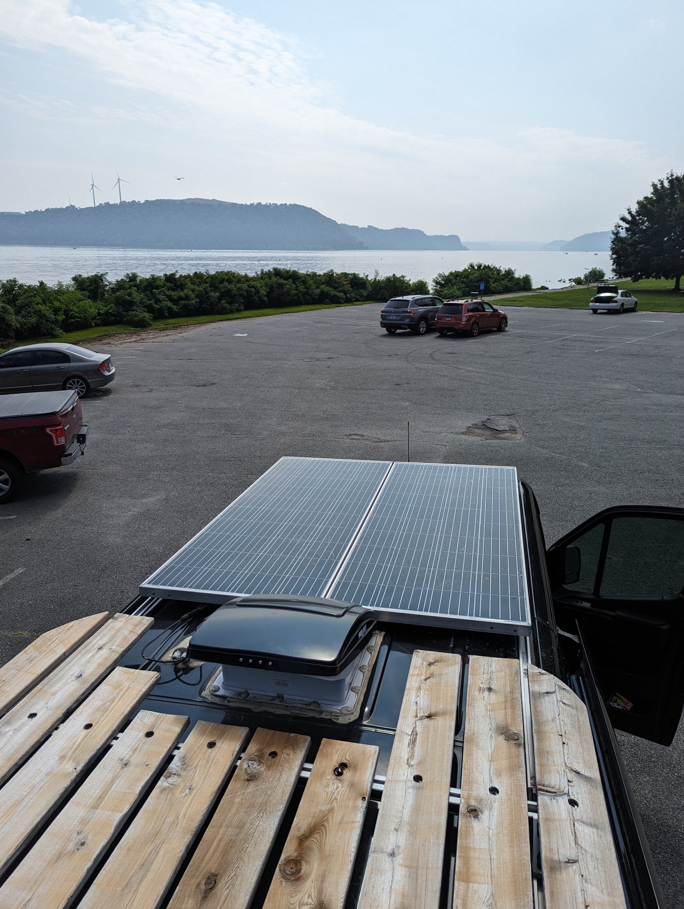 RV rental van rooftop with solar panels & wood rack. Overlooks a parking lot, lake, distant hills, and wind turbines.