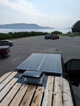 RV rental van rooftop with solar panels & wood rack. Overlooks a parking lot, lake, distant hills, and wind turbines.