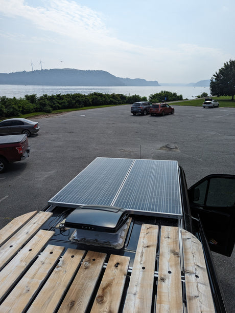 RV rental van rooftop with solar panels & wood rack. Overlooks a parking lot, lake, distant hills, and wind turbines.