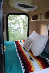 RV rental interior: cozy bed with colorful blanket and window view in a handcrafted Hiker Trailer.
