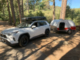 A white SUV with a rooftop tent towing a retro teardrop trailer for RV rental camping fun amidst the trees.