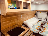 Inside a cozy RV rental: custom wood interior, compact kitchen with cooktop, and a bed with patterned blanket.