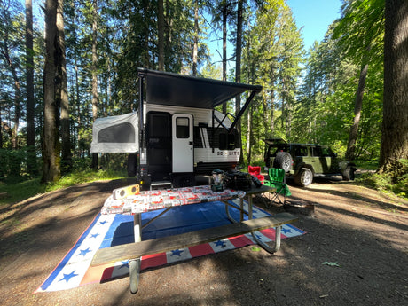 RV rental camping in a sunny forest! A modern camper, olive Jeep, and patriotic picnic setup create an inviting outdoor scene.