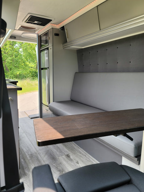 Bright rv rental interior featuring light grey bench seating, dark wood table, fridge, and open door to nature views.