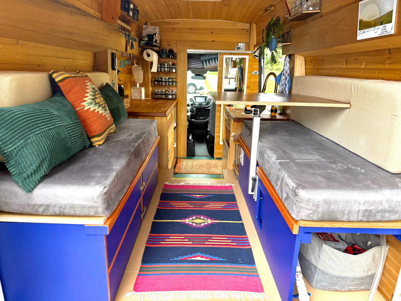 Cozy RV rental interior: wood-lined, vibrant blue storage, grey cushions, colorful pillows, and a compact kitchen. Road trip ready!