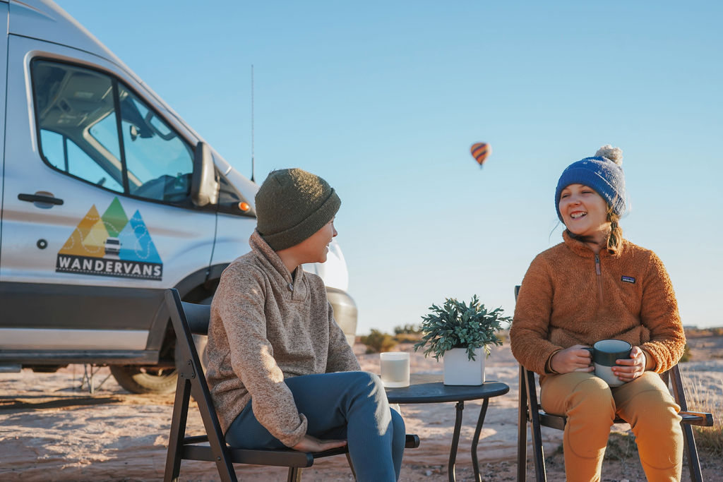  with kids cozy inside and parents taking in the sunset view from the open camper van.|Four friends gather around a crackling campfire beside their Wandervan RV rental — photo 4
