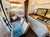 RV rental: Bright camper van interior with bed, kitchen, and open dual-zone portable fridge, parked in desert.