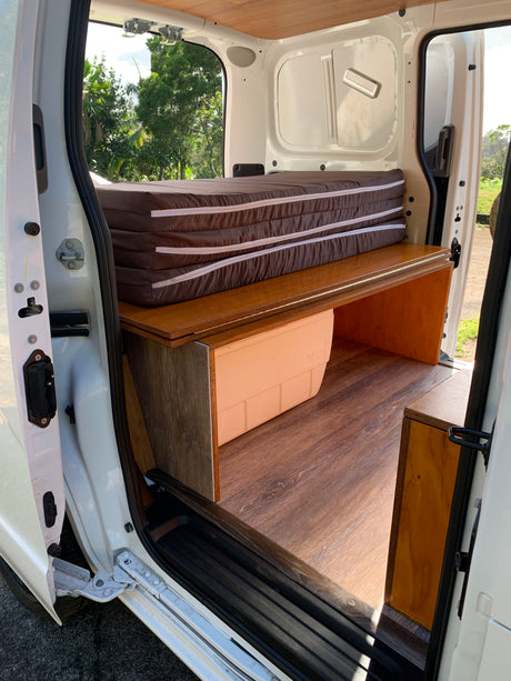 Inside an RV rental camper van: compact bed setup with folded mattress, wood platform, cooler, & storage.