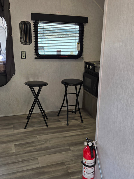 RV rental interior: kitchen with microwave, window showing an outdoor view, two black folding bar stools, and a fire extinguisher.