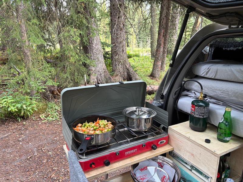 Dinner cooking on the slide-out camp stove at a forest campsite, with the made bed visible through the open hatch — photo 9