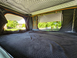 Cozy interior of an RV rental rooftop tent, featuring a quilted sleeping area and open windows with outdoor views.