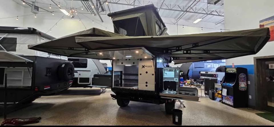 XPedition Voyager Sky XE fully deployed indoors with both Bush Company 270 XT MAX awnings swung out over each side, rooftop tent open on the roof rack, and the rear galley hatch raised over the slide-out camp kitchen — photo 2