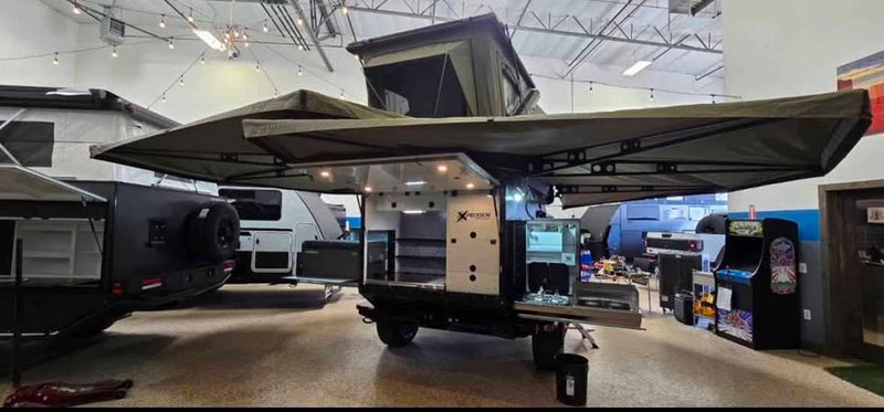 XPedition Voyager Sky XE fully deployed indoors with both Bush Company 270 XT MAX awnings swung out over each side, rooftop tent open on the roof rack, and the rear galley hatch raised over the slide-out camp kitchen — photo 2