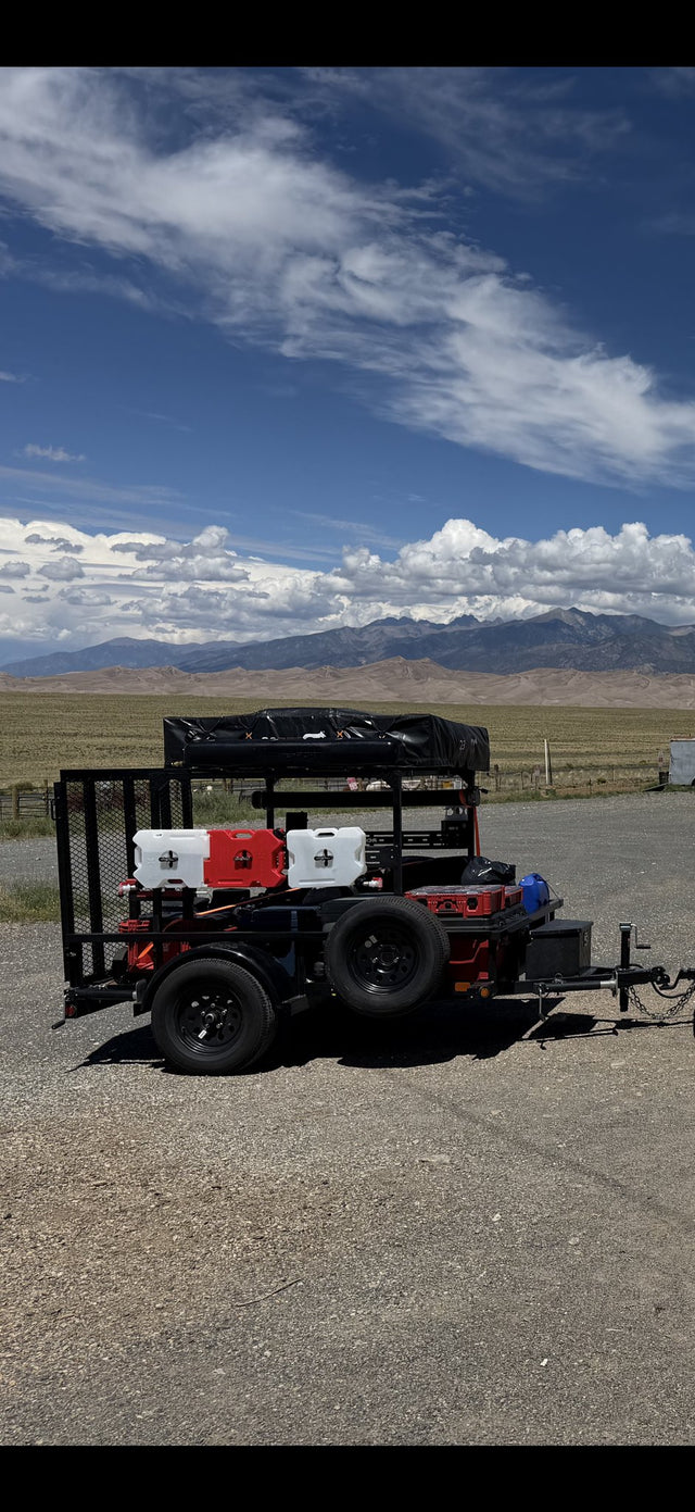Big Tex SA-30 utility trailer broadside on a gravel pullout with the Great Sand Dunes and the Sangre de Cristo range behind it, showing the custom elevated square-tube steel RTT platform carrying the covered 23ZERO rooftop tent, the mesh ramp gate, spare tire, tongue box and fuel cans mounted along the side rail