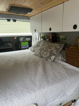 Inside a comfy RV rental: bed with grey bedding, wood ceiling, overhead cabinets, and a view outside the window.