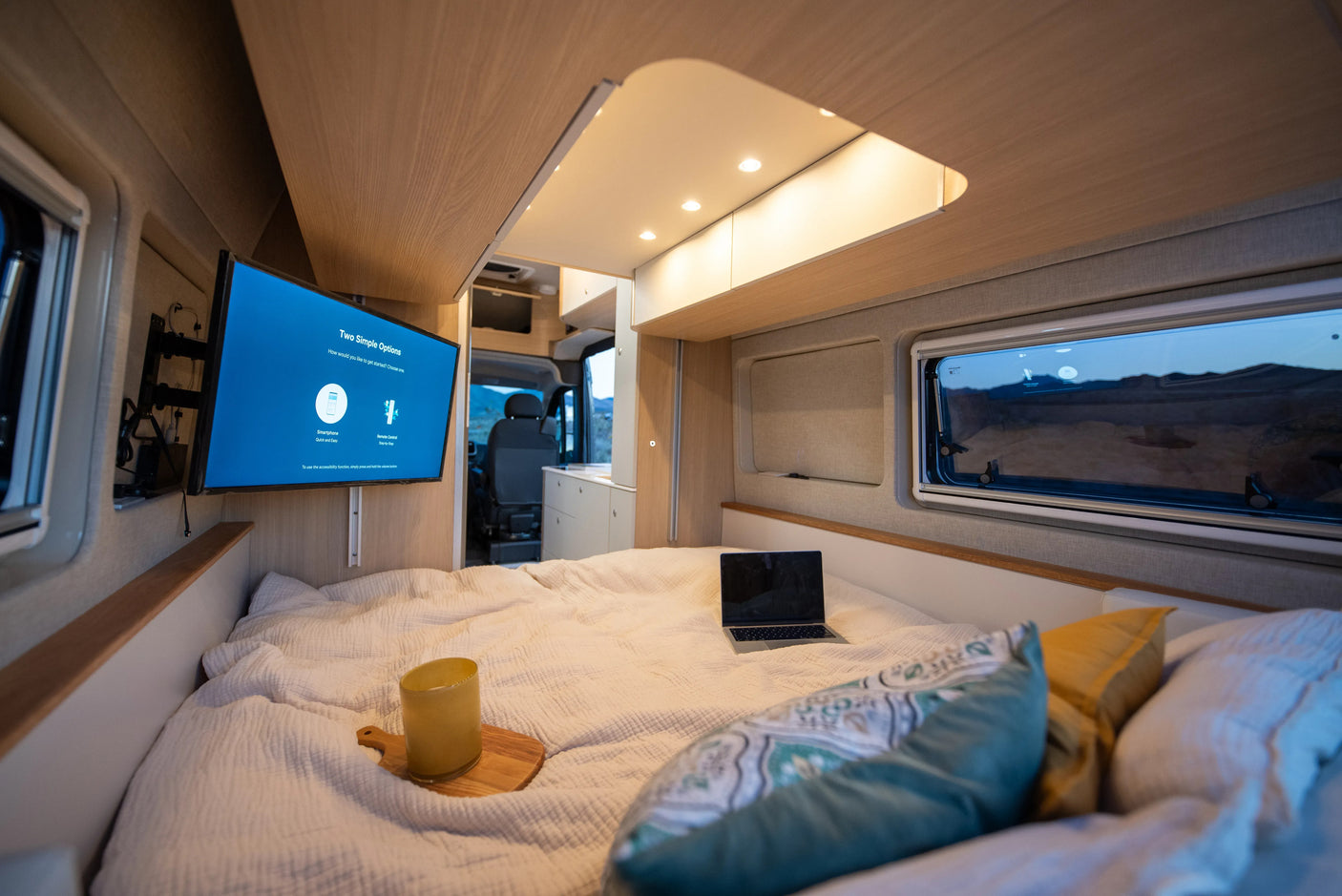 Cozy RV rental bedroom: TV showing setup options, laptop on bed, and window view of a serene landscape.