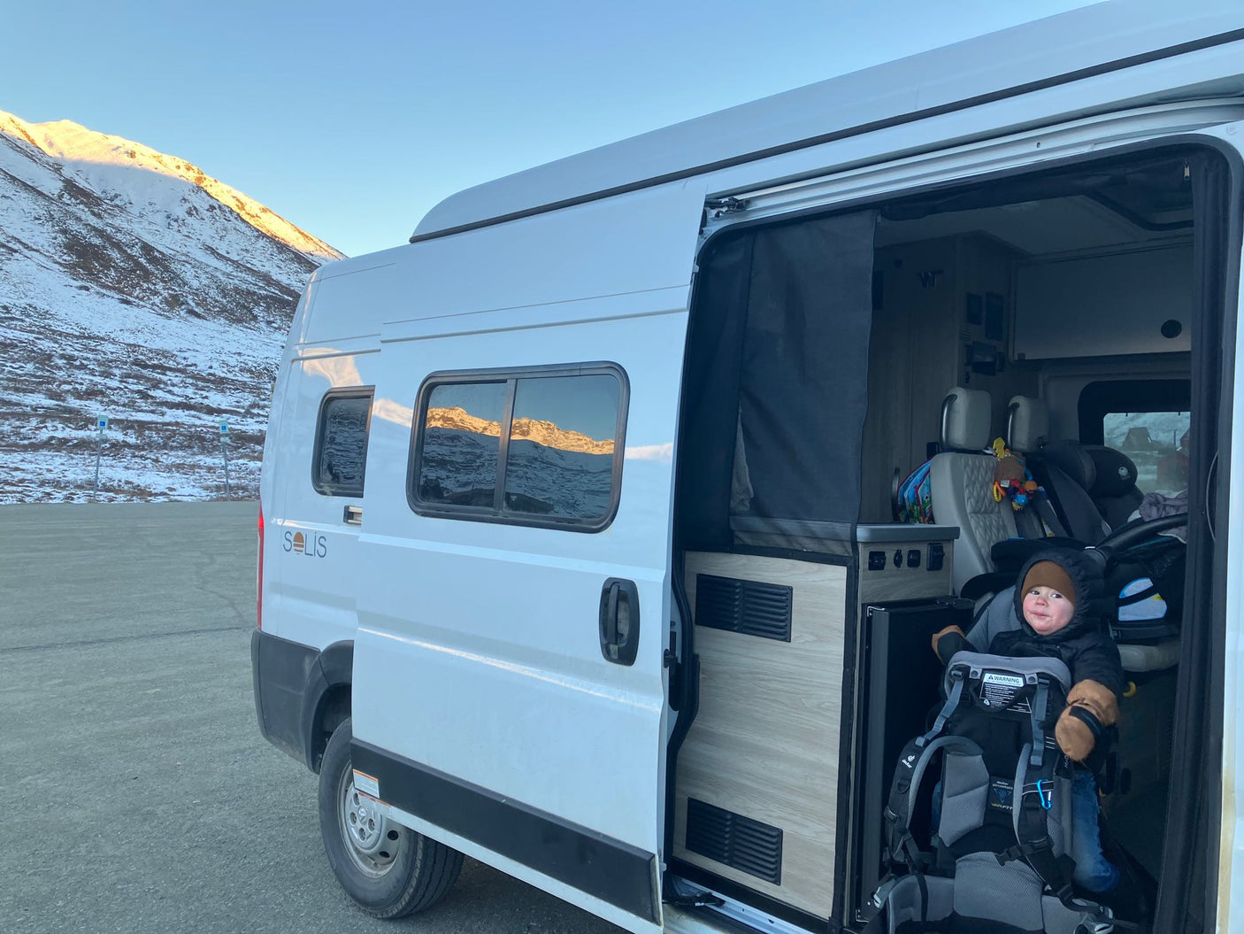 Winter RV rental trip: A happy baby in a carrier inside a Solis camper van, parked amidst snowy mountains.