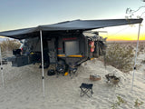 An off-road camper trailer, ideal for RV rental, with an awning & kitchen set up on sandy terrain at sunset.