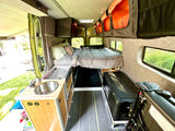 RV rental van interior features a cozy bed, kitchenette with sink/stove, and overhead storage for adventurers.